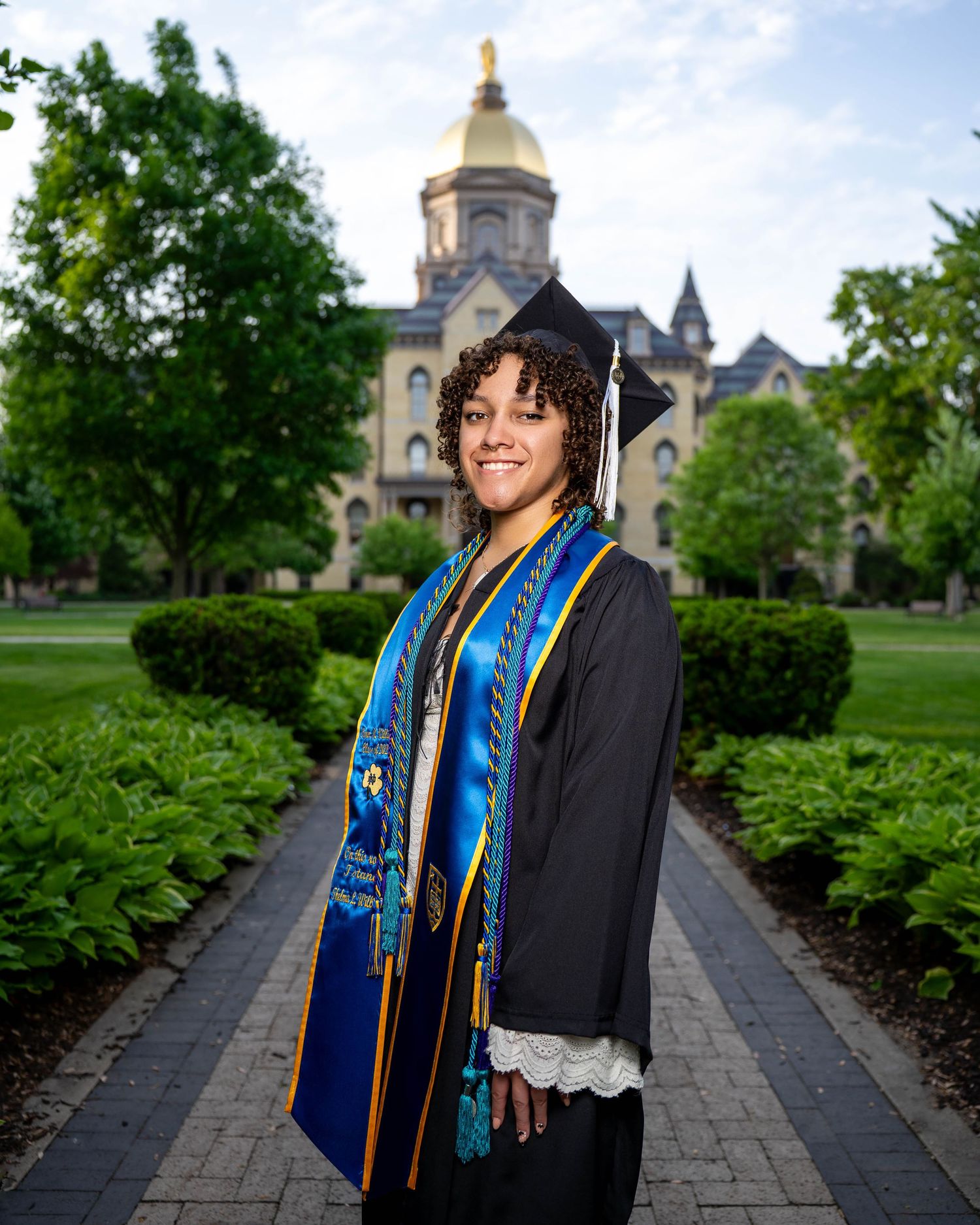 A graduate in cap and gown stands on a pathway with Notre Dame's Golden Dome visible in the background.