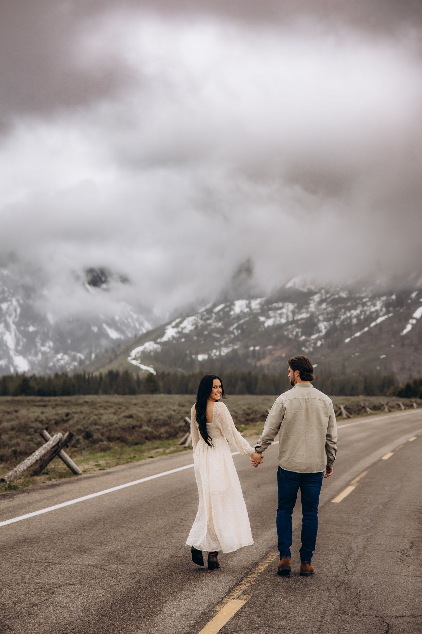 A couple holding hands walks down an empty mountain road with dramatic cloudy peaks in Grand Teton National Park.