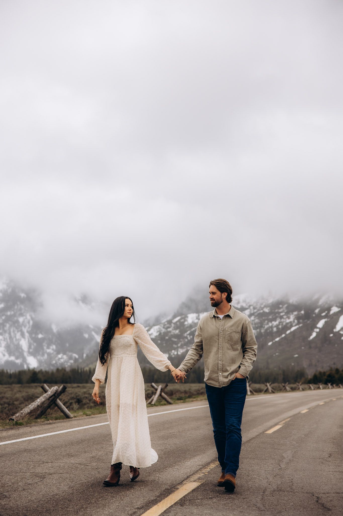 A couple walks hand in hand on a mountain road with misty peaks and cloudy skies in the background.
