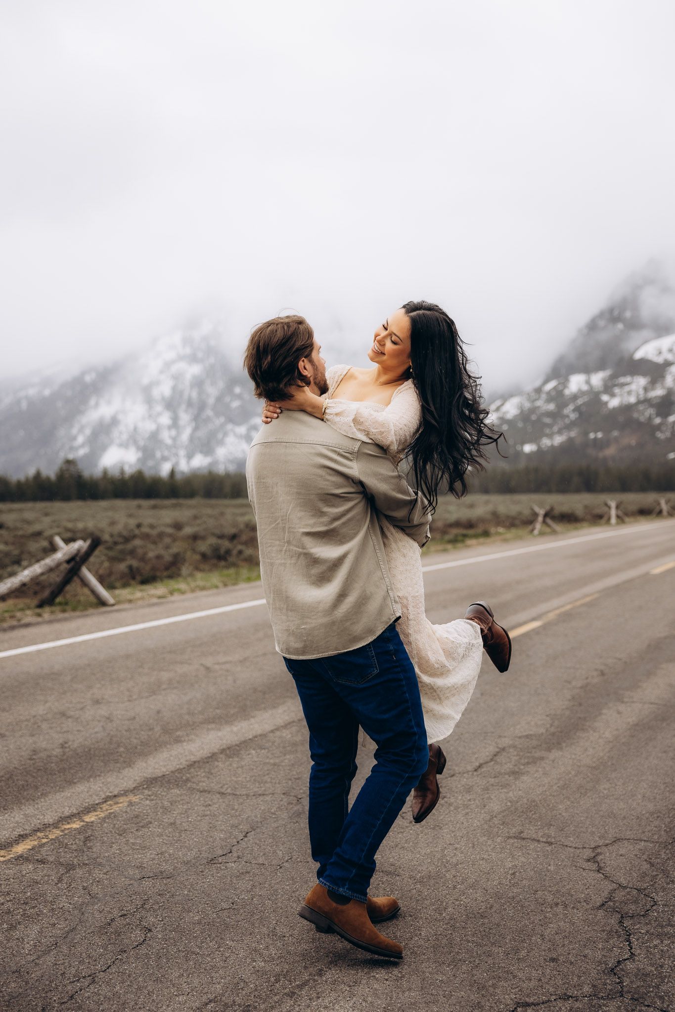 A couple embraces on a mountain road with snow-capped peaks and foggy mountains in the background near a rural highway.