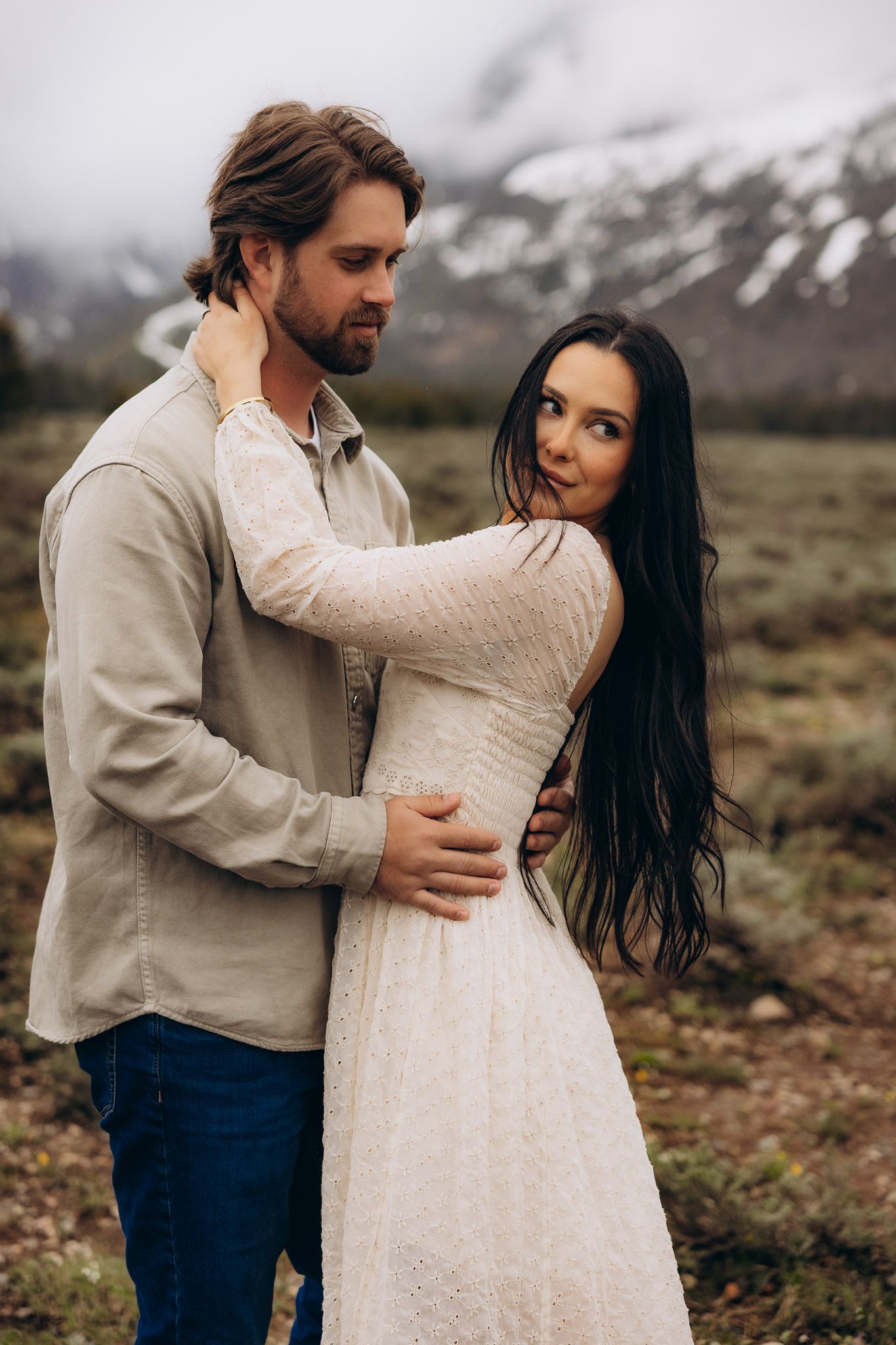 A romantic couple embraces against a backdrop of misty mountains, wearing white and beige outfits in a rustic outdoor setting.