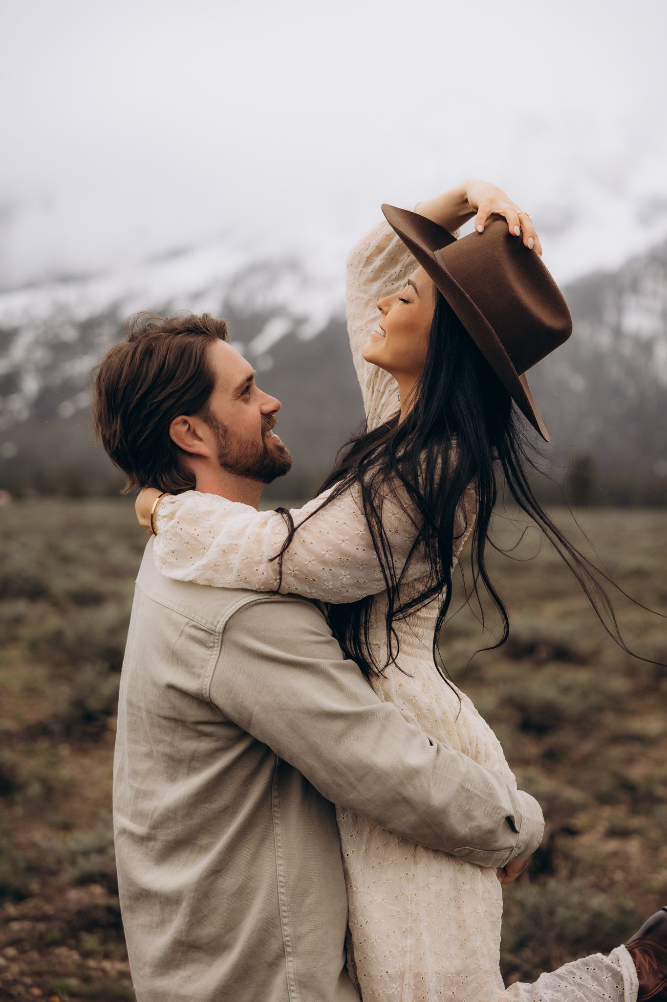 Romantic couple embraces in casual attire against snowy mountain backdrop as woman tilts head back wearing brown hat.
