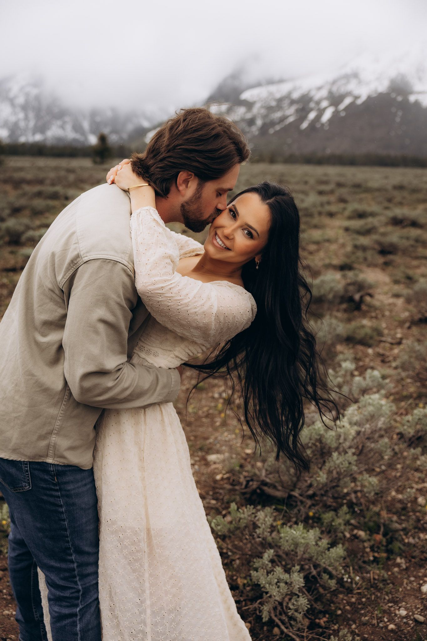 A romantic couple shares an intimate moment in the wilderness with snow-capped mountains visible in the background.