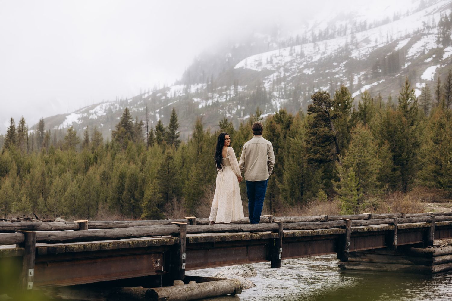 A couple stands on a wooden dock surrounded by evergreen forest and snow-capped mountains in a serene lake setting.