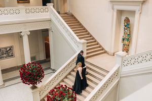 Person in dark dress walking up elegant white marble staircase with ornate railings and red floral arrangements.