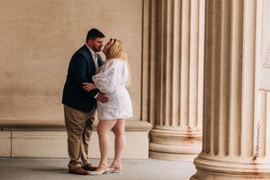 A romantic couple stands between classical stone columns during an engagement photo session.
