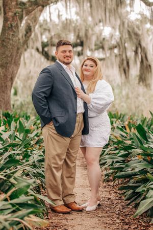 A couple poses together on a garden path surrounded by lush greenery and Spanish moss hanging from trees in the background.