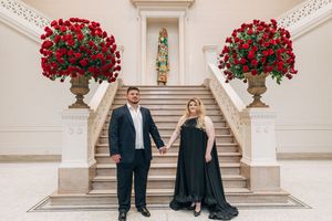 A couple stands on elegant white stairs flanked by large decorative urns filled with vibrant red roses.