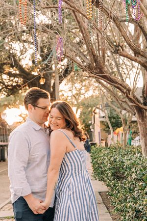 A couple embraces outdoors on a path with decorated trees and string lights in the background.