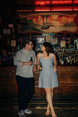 A couple sharing drinks and conversation at a dimly lit vintage bar with red neon lighting overhead.