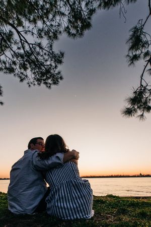 Couple sits together watching sunset over calm waterfront as pine branches frame romantic silhouette against dusky sky.