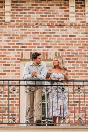 A couple stands together on an ornate wrought iron balcony against a brick wall background.
