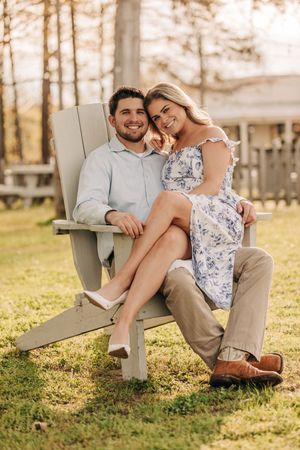A couple sits together in an Adirondack chair on a lawn during golden hour with trees in the background.