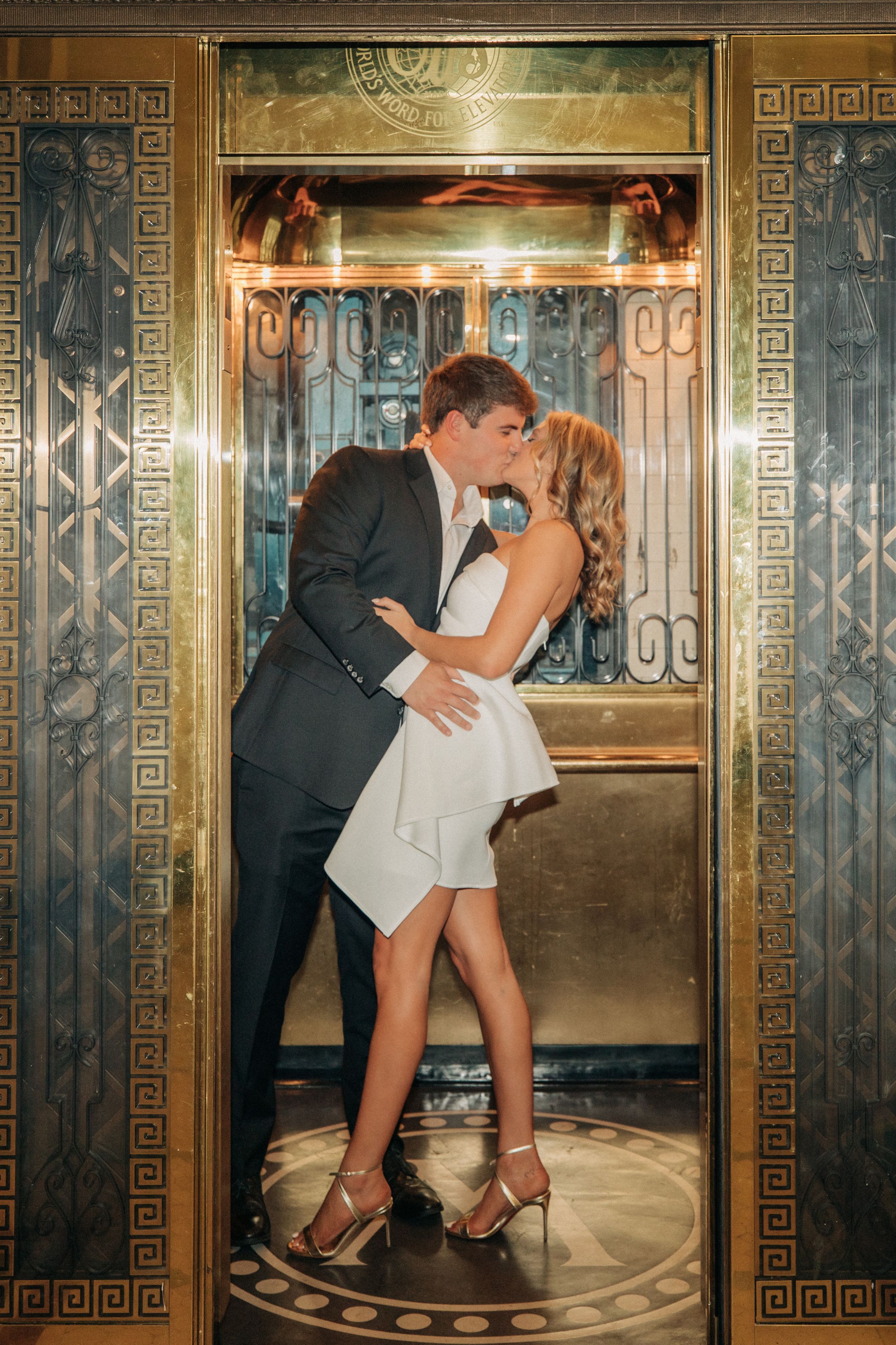 A romantic couple are embracing in a vintage Art Deco elevator with ornate metalwork and geometric patterns.