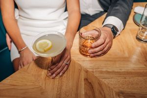 Hand holding a cocktail glass at a wooden bar with multiple beverage glasses visible in sequence.