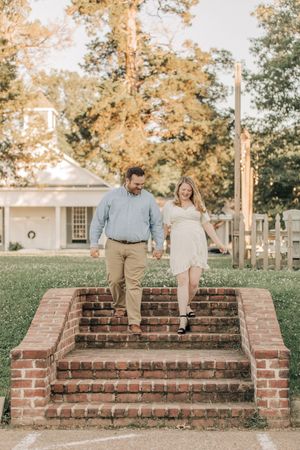 A couple descends brick steps together in an outdoor garden setting with autumn trees in the background.