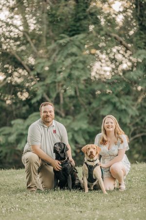 A couple sits on grass with their black and golden dogs during an outdoor photo session.