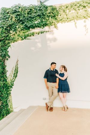 A couple stands together in a romantic setting with white walls and green ivy creating an elegant arch overhead.