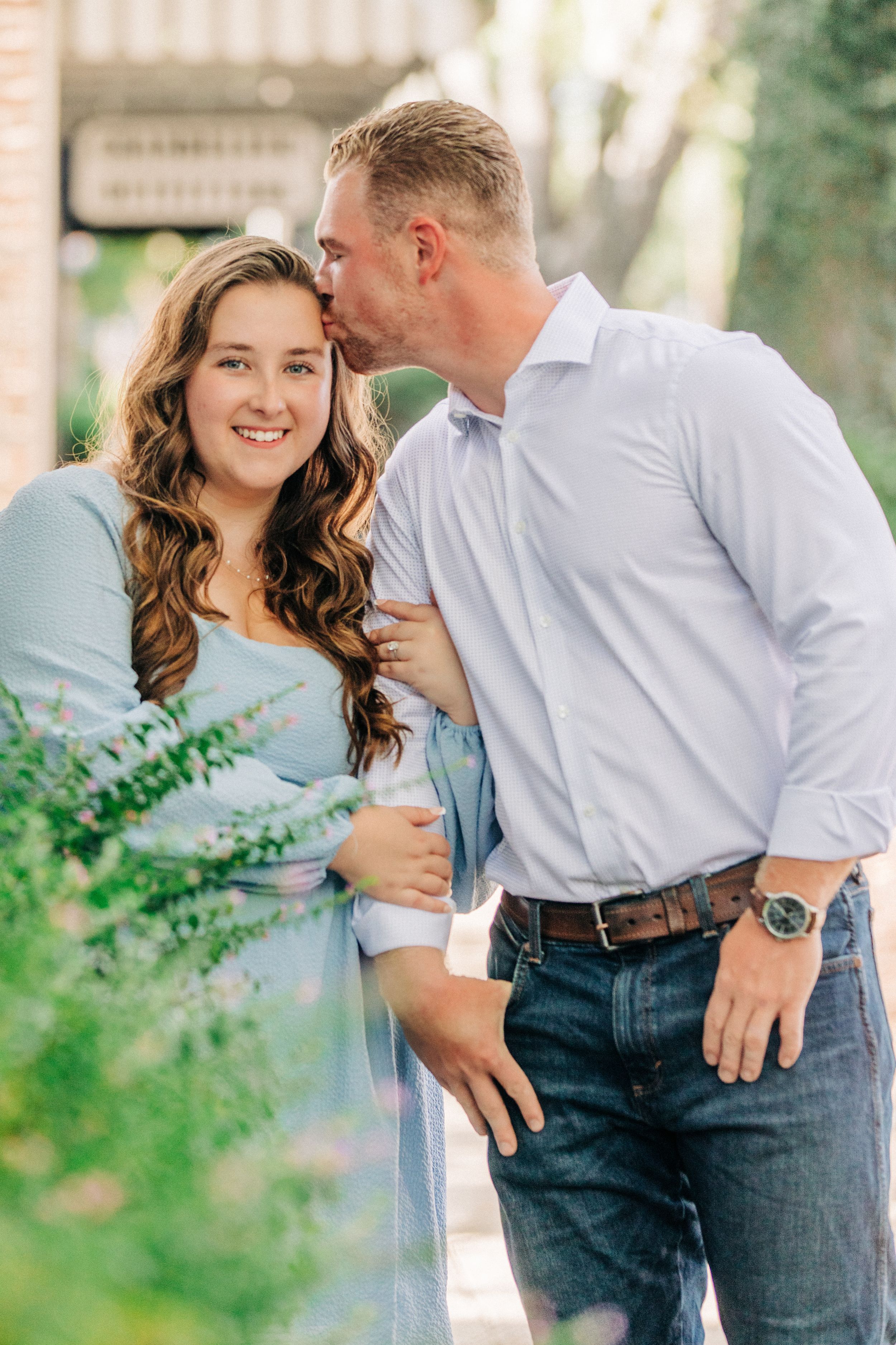 Couple shares intimate moment outdoors among green foliage while wearing casual blue and white clothing.