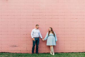 A couple stands together holding hands against a pink brick wall as they smile and gaze at each other.