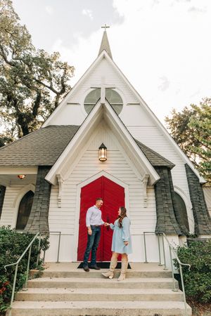 A charming white chapel with a red door and stone accents features a steep A-frame roof and lantern light fixture.
