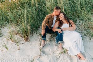 Two people in casual attire sitting close together on sandy beach dunes surrounded by tall grass.