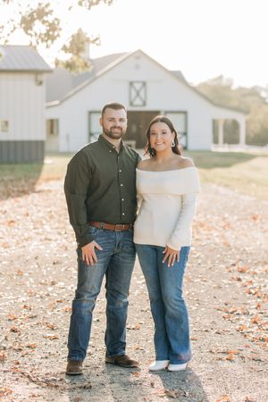 A couple stands together on a gravel path in front of a white barn on an autumn day.