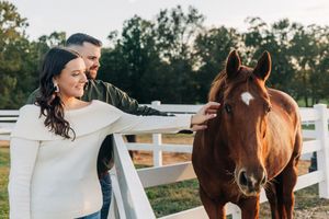 A couple stands next to a beautiful brown horse by a white fence at sunset in a rustic farm setting.