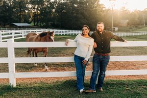 A couple sits together on grassy lawn beside a white fence during sunset creating a romantic outdoor scene.