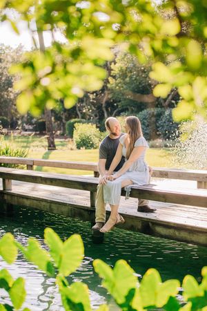 Couple sits together on wooden dock over tranquil pond with green foliage framing romantic lakeside scene.