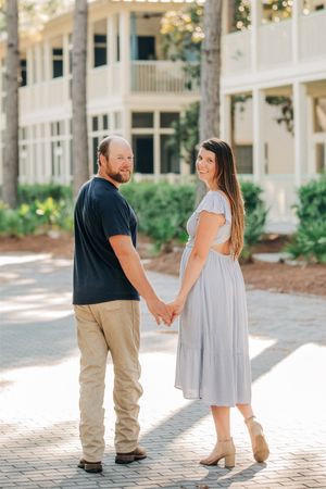 A couple holding hands while standing on a brick path in front of a white colonial-style building on a sunny day.