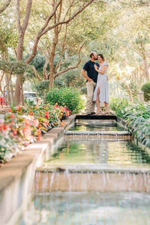 A couple shares a romantic moment on a wooden bridge over cascading water features in a lush garden setting.