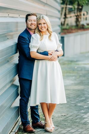 A couple in formal attire poses against a gray wall outdoors, with the person in a navy suit embracing their partner in white.