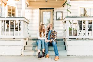 A couple sits relaxing on white painted porch steps of a charming vintage farmhouse on a sunny afternoon.