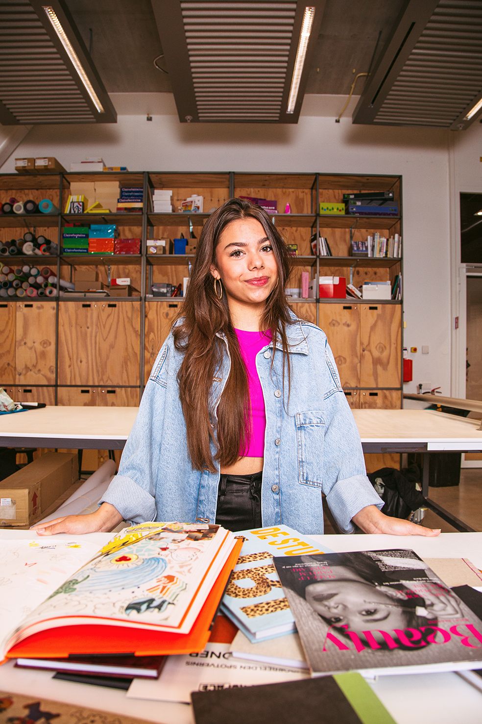 Student in denim jacket studying with books and magazines spread across a wooden table.