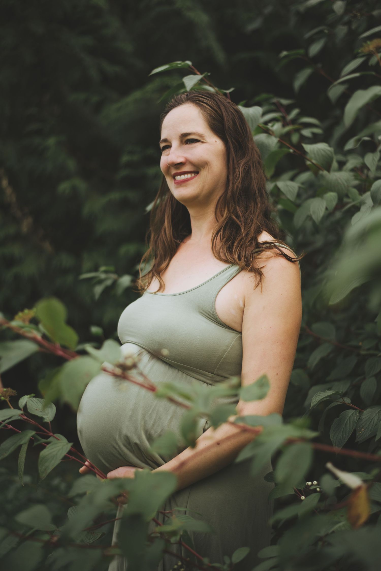 An expectant mother in sage green dress smiles while standing among leafy foliage.