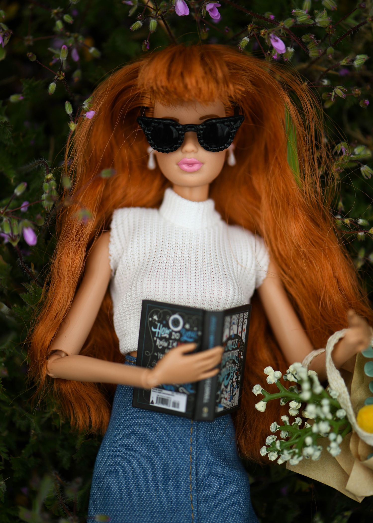 Red-haired Barbie doll wearing sunglasses, white knit top, denim skirt, holding a book and floral tote bag outdoors.