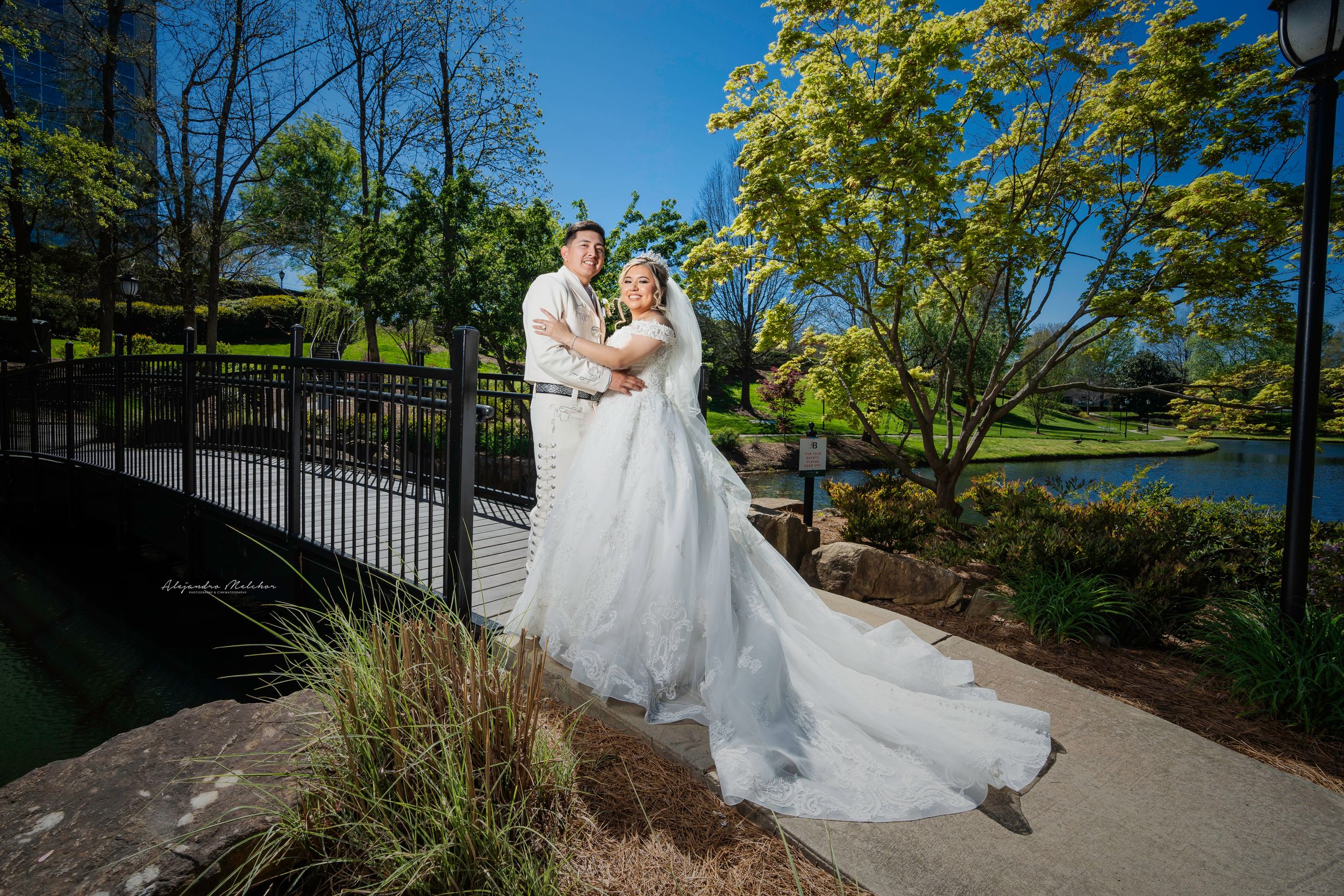 A wedding couple poses on a bridge over a pond surrounded by autumn foliage and landscaped gardens.