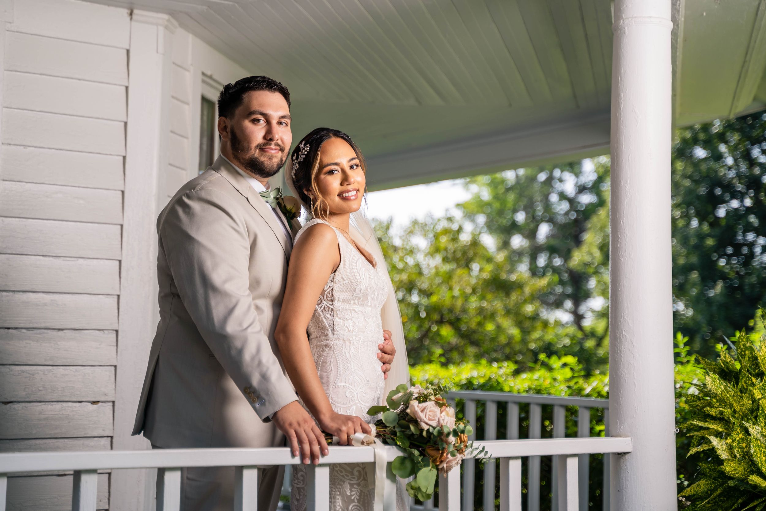 Elegant couple in wedding attire pose together on a white porch during golden hour.