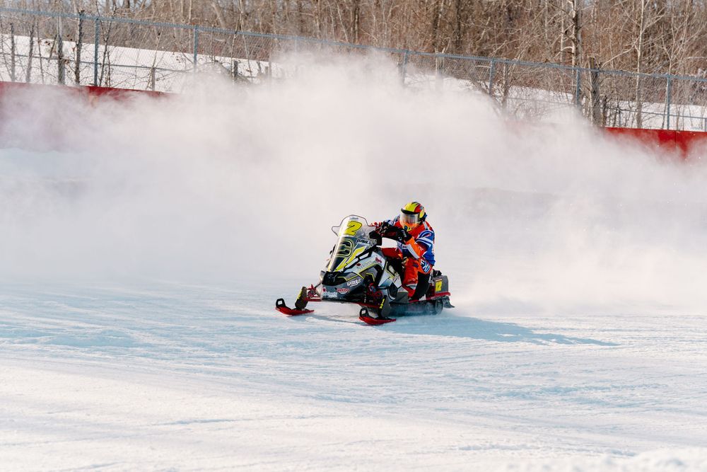 A snowmobile racer speeds through a snowy track, kicking up a cloud of snow dust during a winter racing competition.
