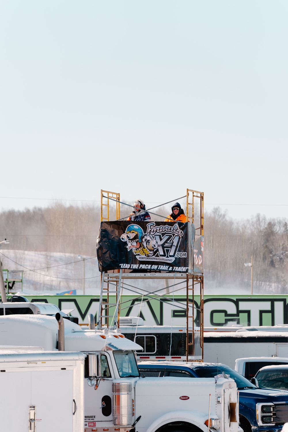 Two people stand on a scaffolding tower with a banner at a snowy outdoor racing event surrounded by trucks.