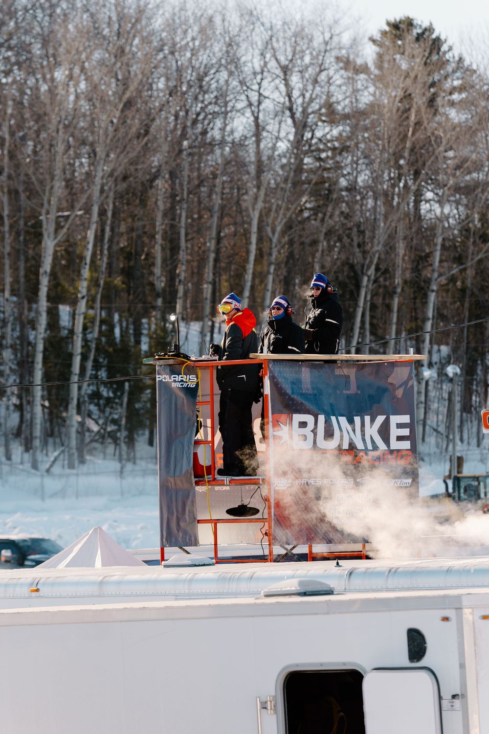 Officials stand on an elevated platform at a winter snowmobile racing event with Polaris Bunke signage.