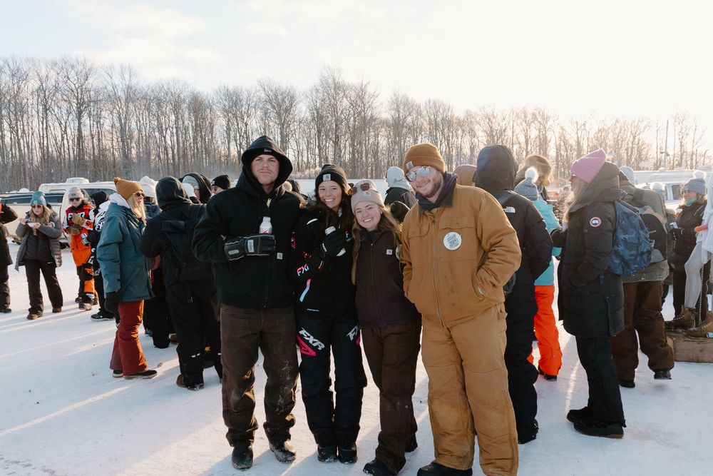Group of friends posing outdoors at a winter snowboarding event with snowy landscape and trees in background.