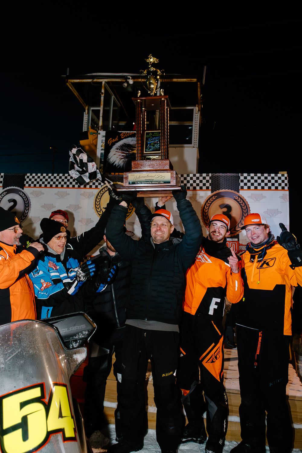 Team celebrates snowmobile racing victory holding trophy at night finish line with checkered flag.