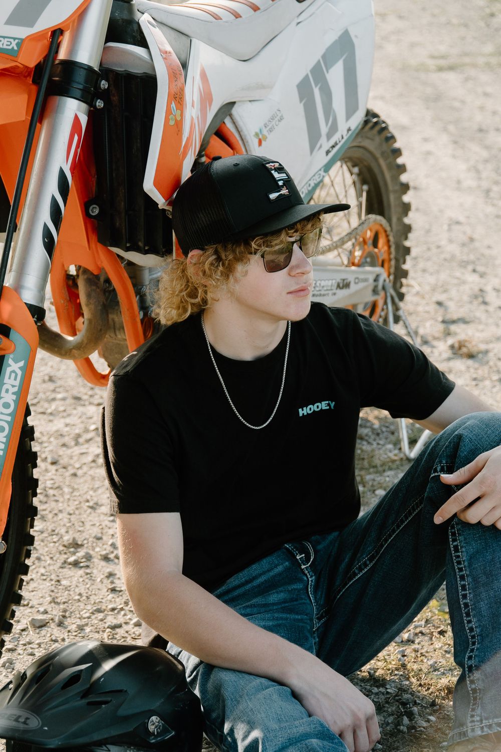 Person in black t-shirt and cap relaxes against orange motocross bike outdoors.