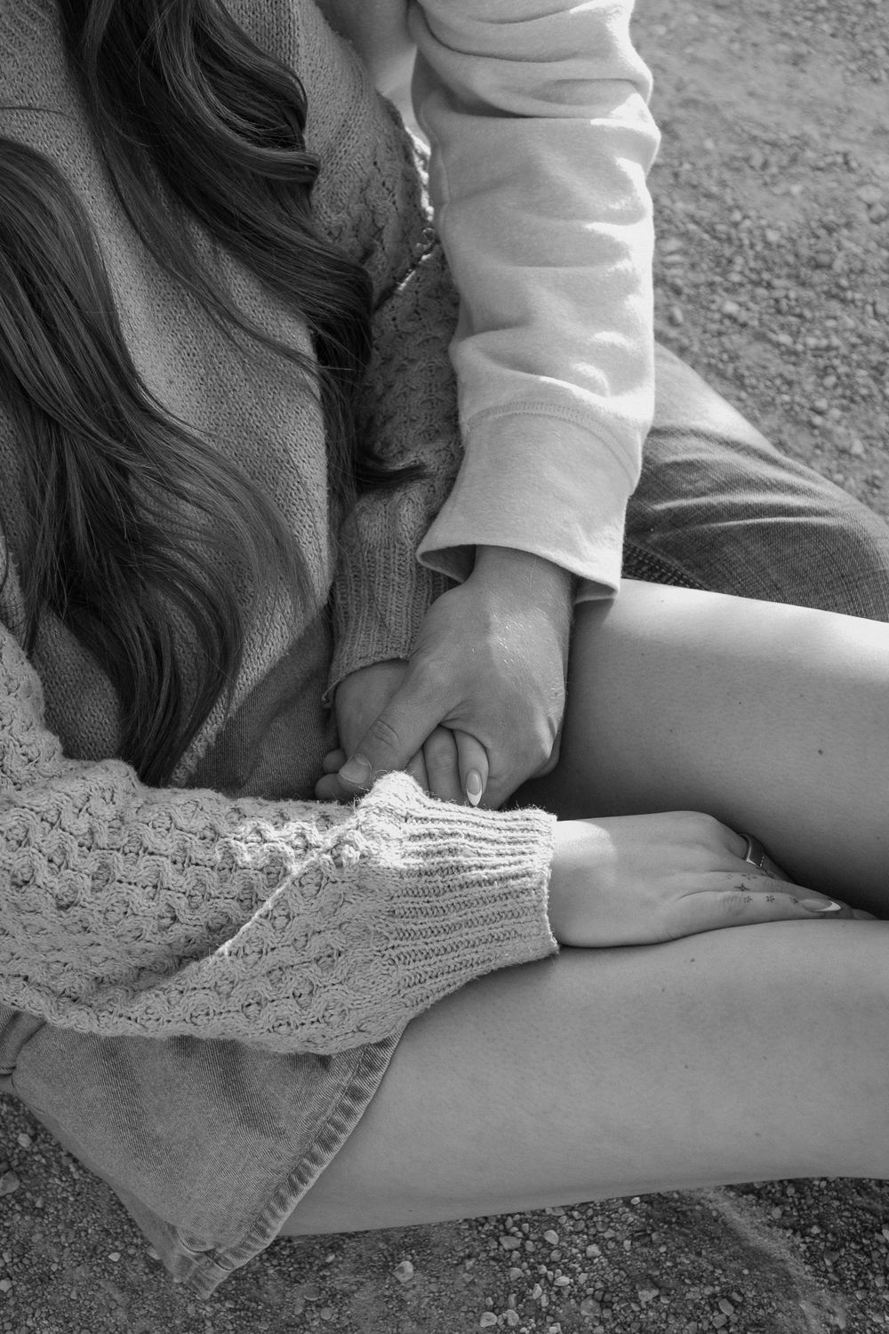 Black and white photo of a couple holding hands while sitting outdoors on gravel ground.