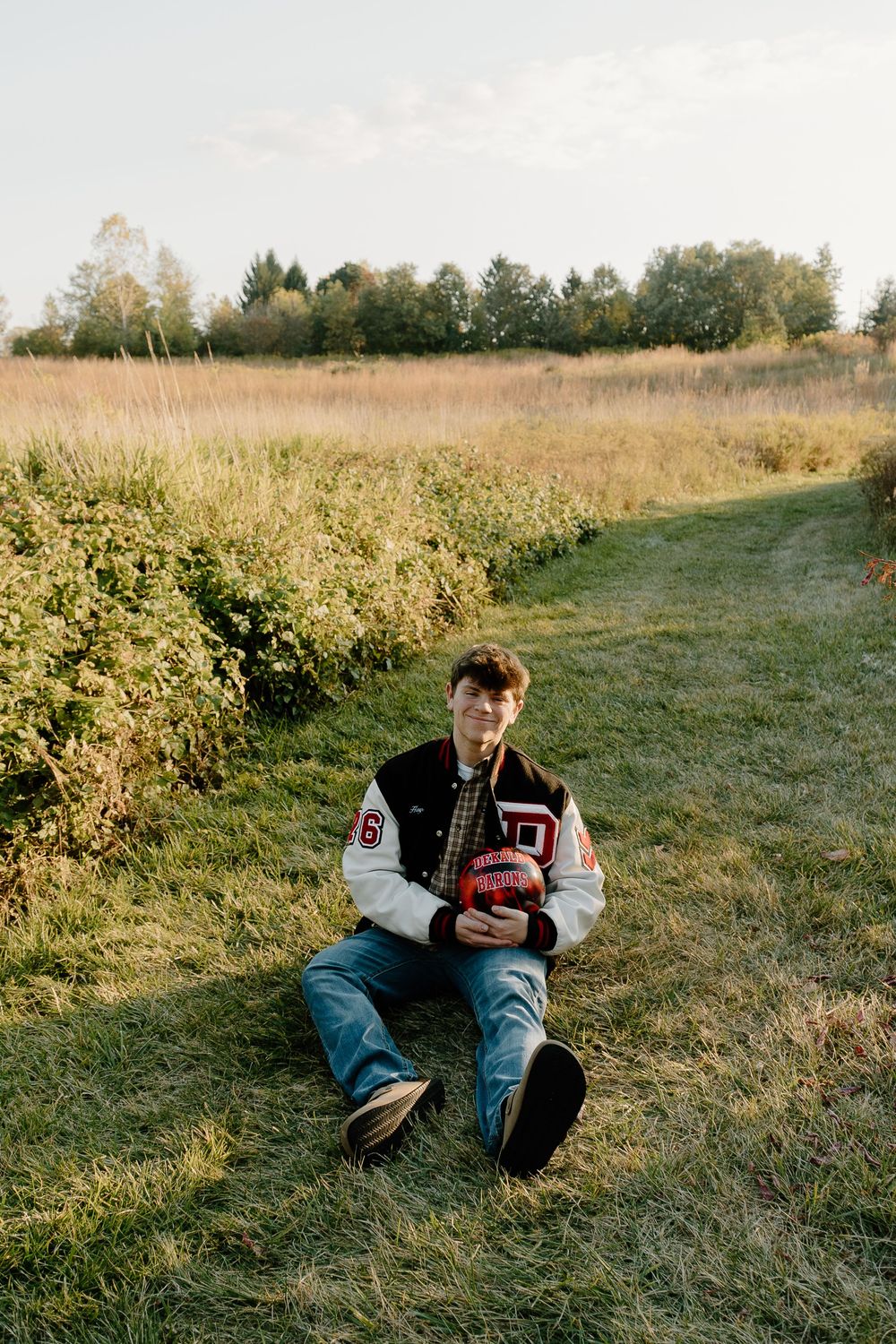 A young man in a varsity jacket and jeans sits on a grassy path in a rural field holding a football.