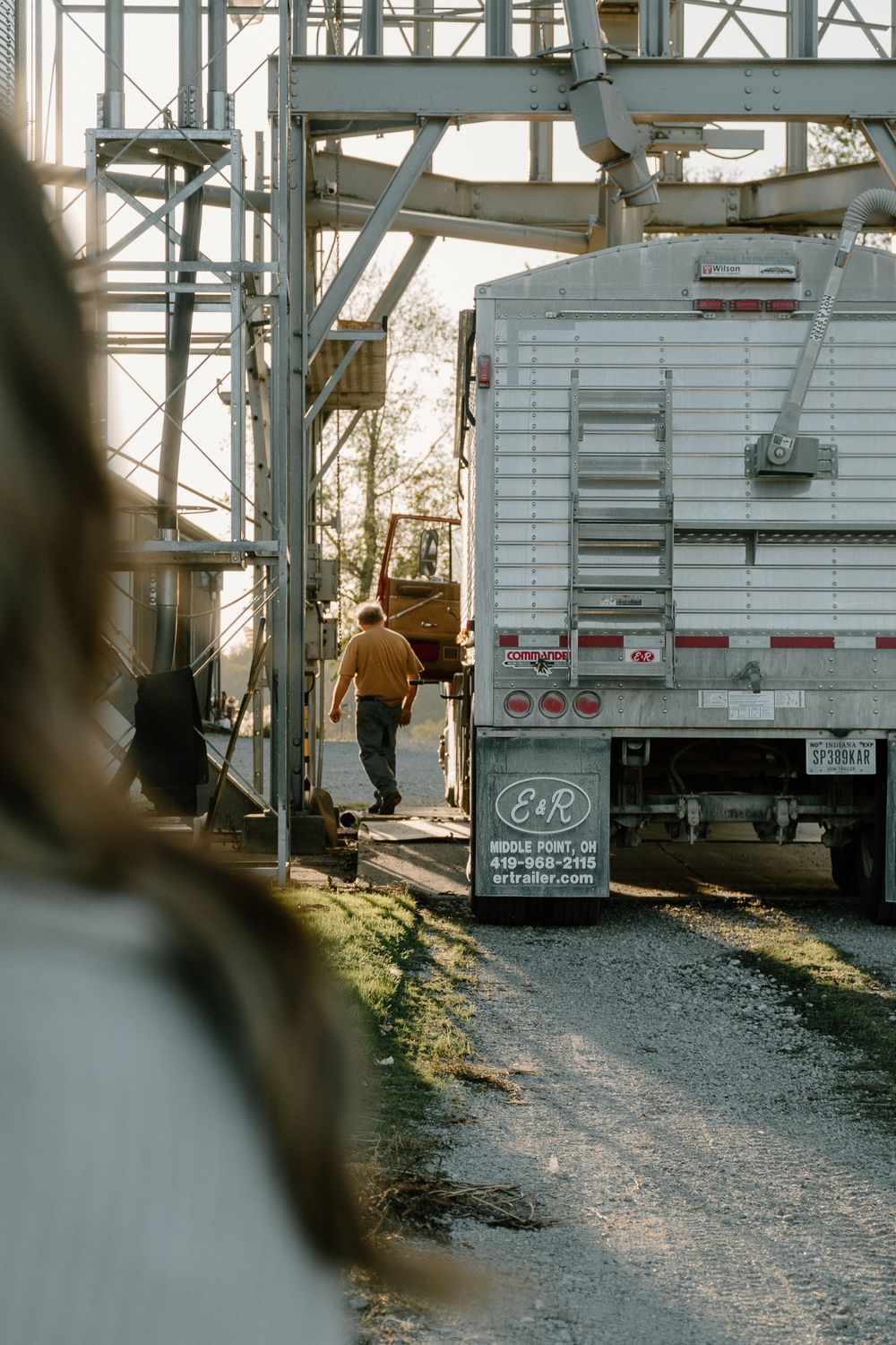 Worker climbing industrial equipment near transport truck in late afternoon light.