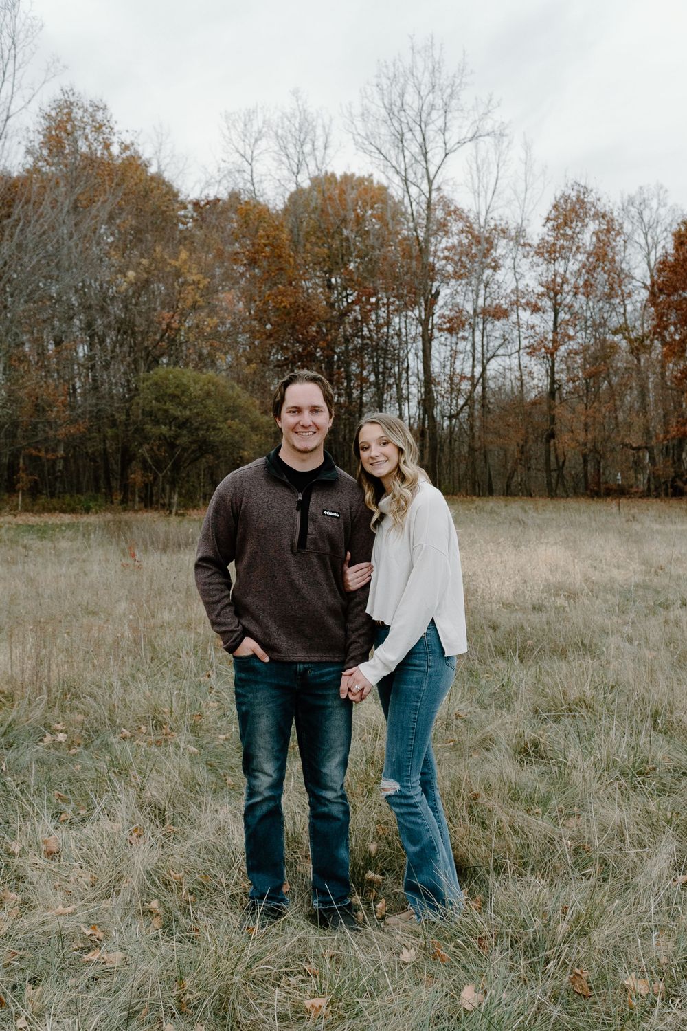 A couple stands together in a rustic autumn field with colorful trees in the background.