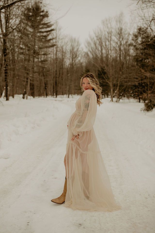 A woman in a flowing white dress poses on a snowy winter path surrounded by evergreen trees.